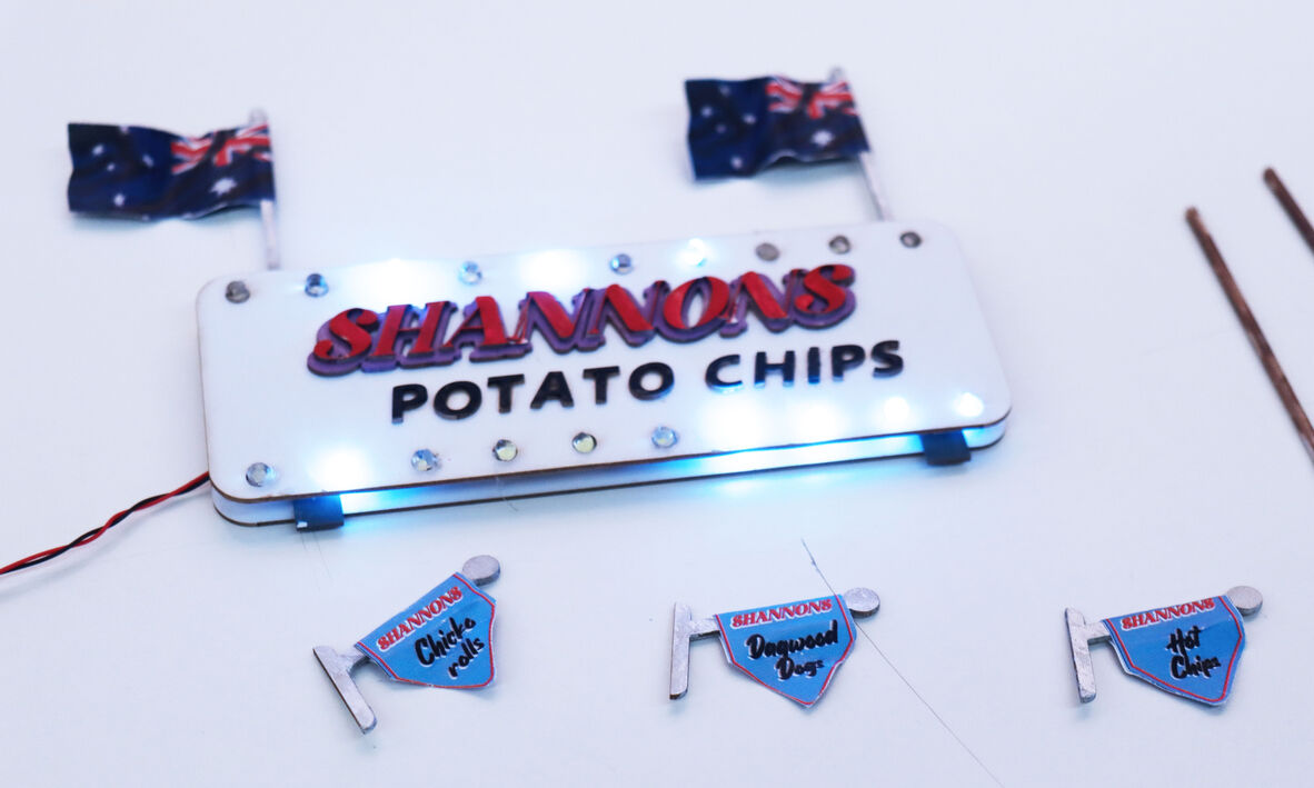 A retro-inspired illuminated sign with the text "SHANNONS POTATO CHIPS" in bold red and black letters, adorned with small lights along the edges. Two small Australian flags are displayed above the sign. Below the sign are three mini flags labeled with different snack items: "Chiko Rolls," "Dagwood Dogs," and "Hot Chips."