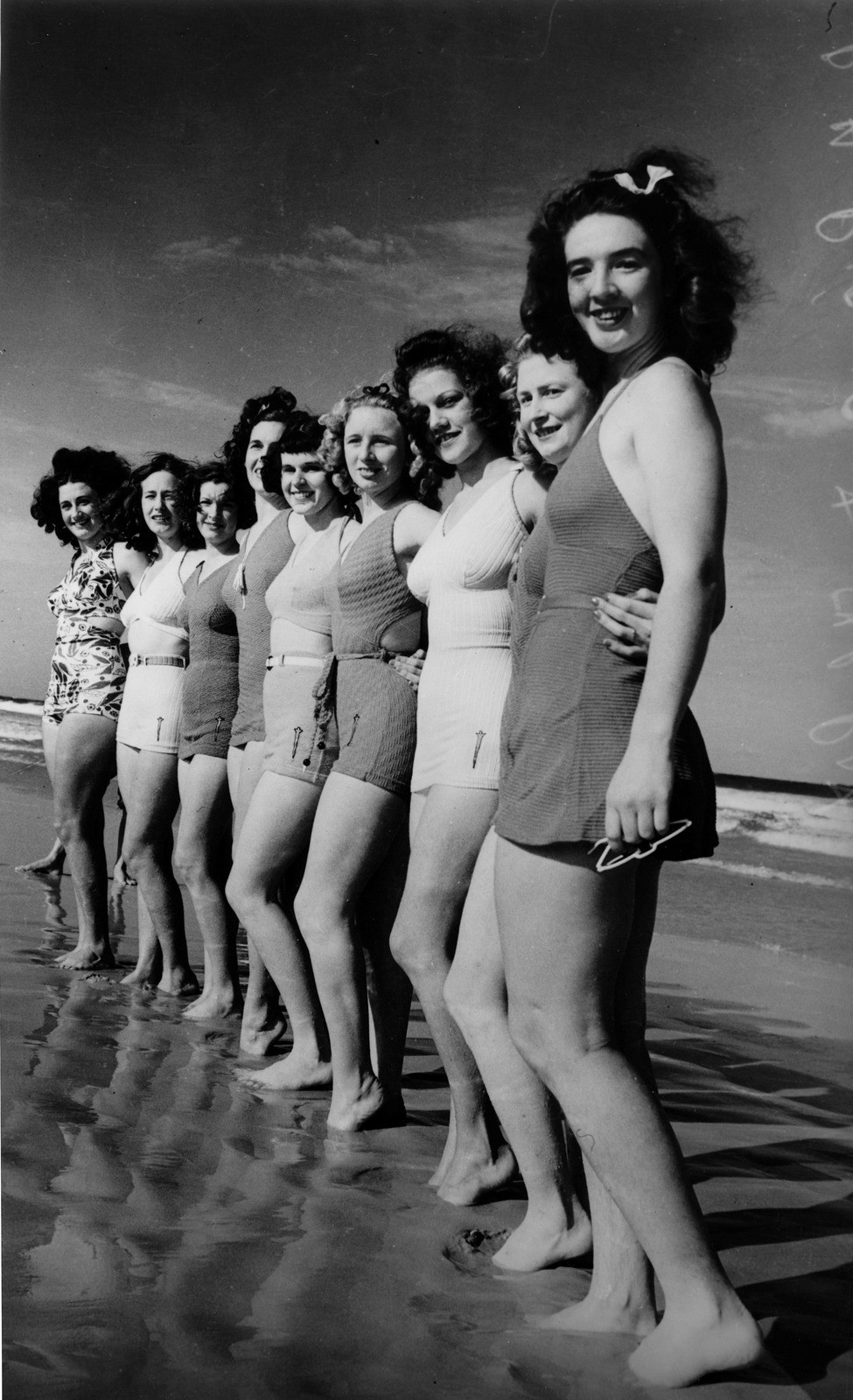 Photo of ten women posing in a line in bathing suits at the edge of the water at a beach in 1939