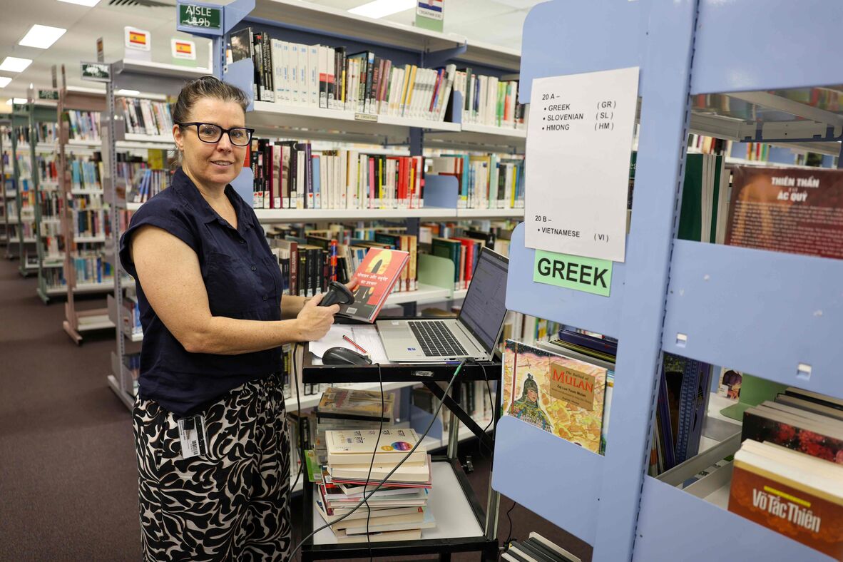 Woman checking out a book via computer on a library trolley next to a shelf labelled Greek