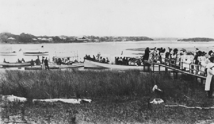 Black and white image of passengers standing on a wooden jetty waiting to board small boats in the Maroochy River. Some passengers are sitting in a boat about to leave the jetty. ca.1918