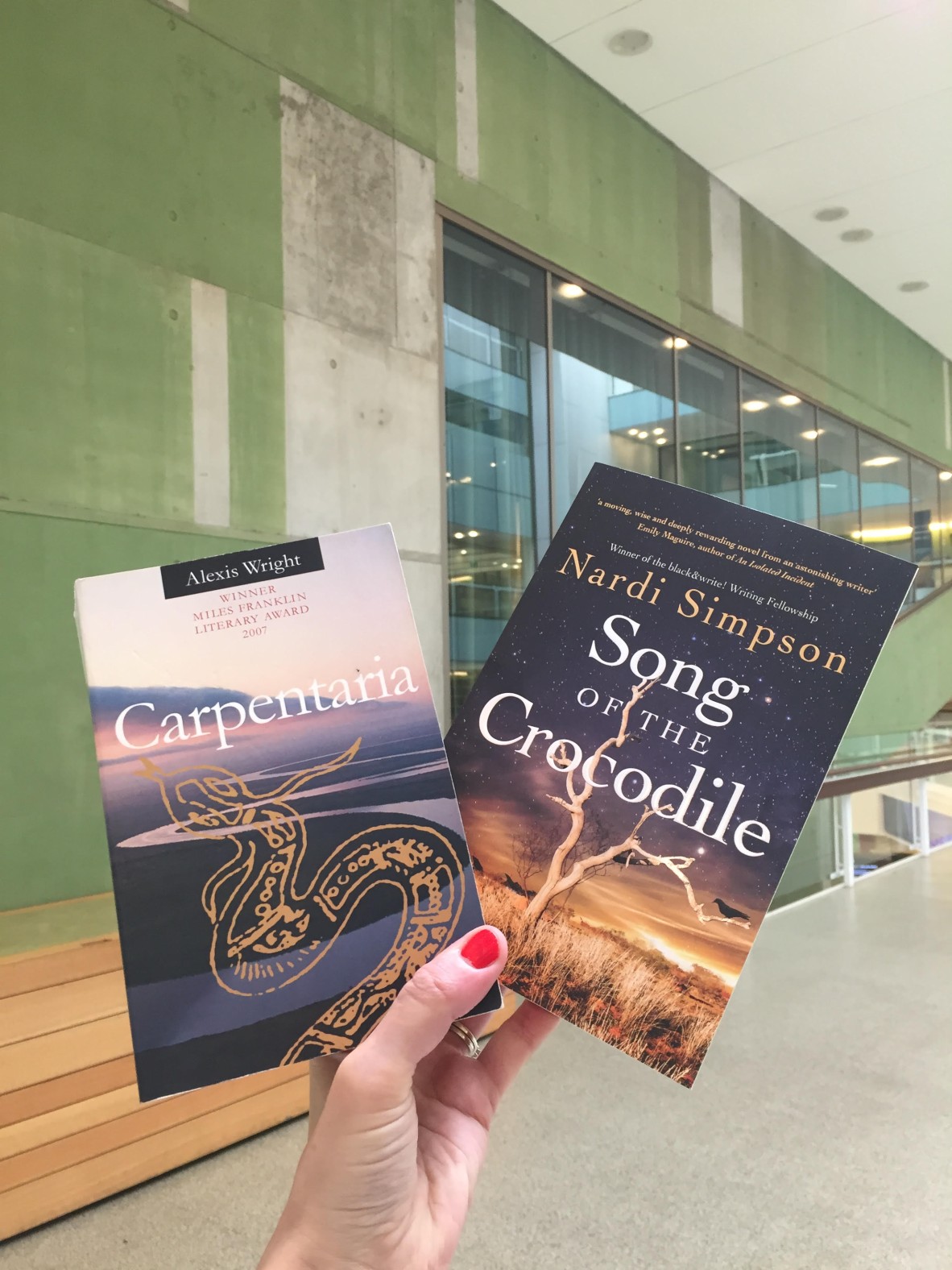 A hand holds up 'Carpentaria' by Alexis Wright and 'Song of the Crocodile' by Nardi Simpson against a wall at State Library