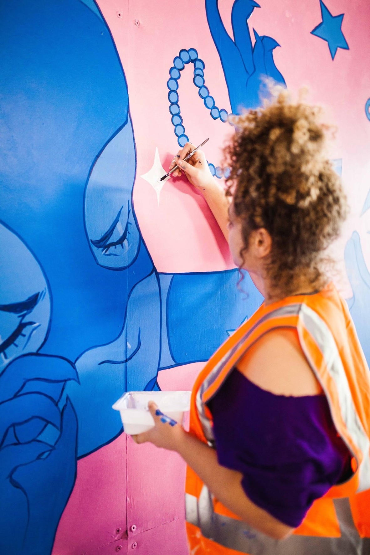 Tori-Jay Mordey wears a high vis vest and a purple shirt She is standing side on painting a blue and pink mural