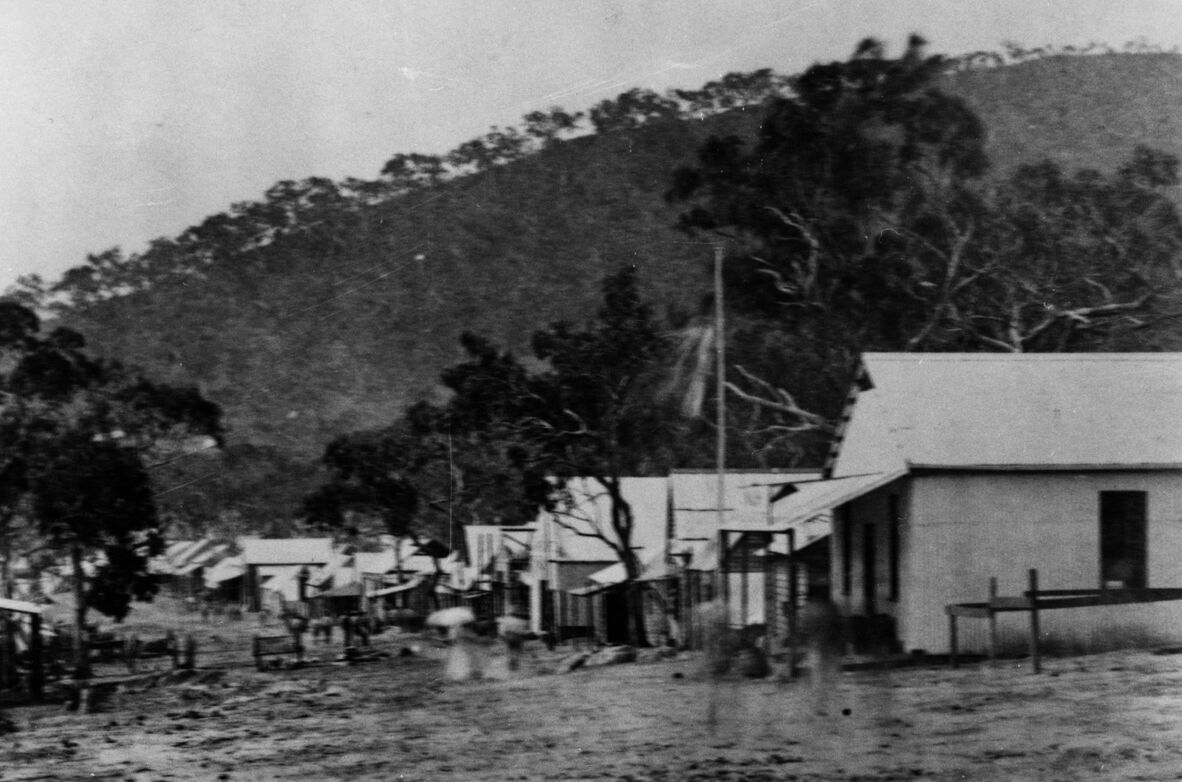 A black and white photo of a town with small huts and houses and a mountain in the background