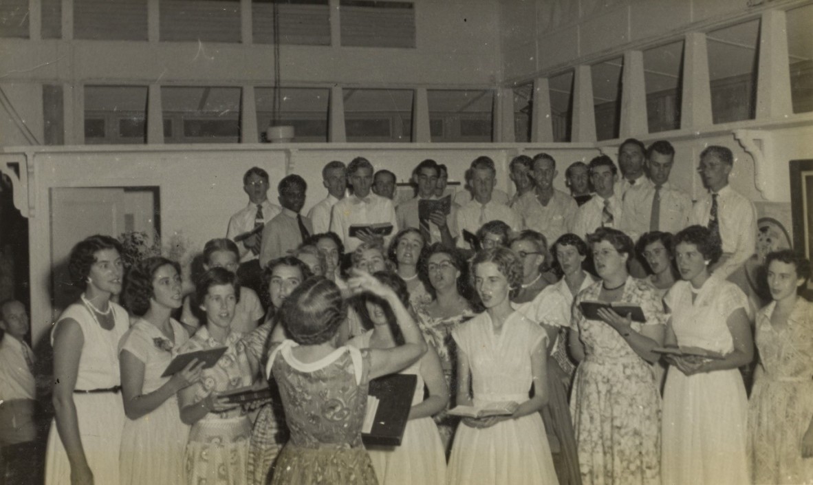 Sepia photo of group of people standing in five rows singing 1955