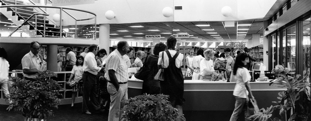 Black and white photograph showing a busy library circulation desk with patrons and plants in the foreground