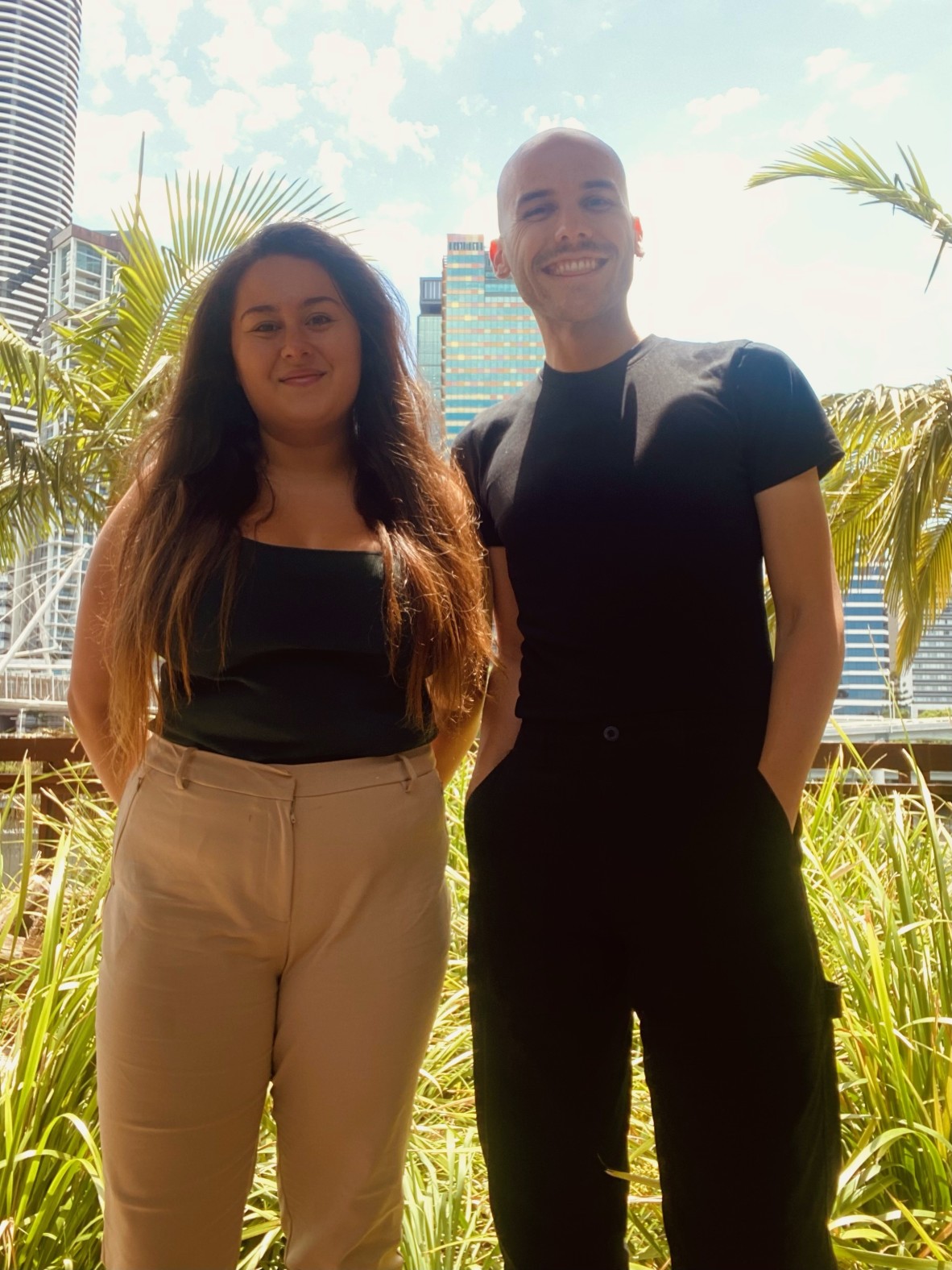 Mid shot of Georgia and Darby standing with buildings and plants in the background. The sun is shining and both are smiling at the camera.