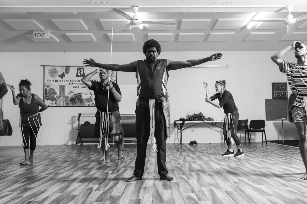 Deaf Indigenous Dance Group dancers rehearsing in Cairns, 2021. A dancer stand in the foreground with their arms outstreched. The background dancers are wearing string skirts over their normal clothing. The men have fur attached to their skirts and are holding prop spears.