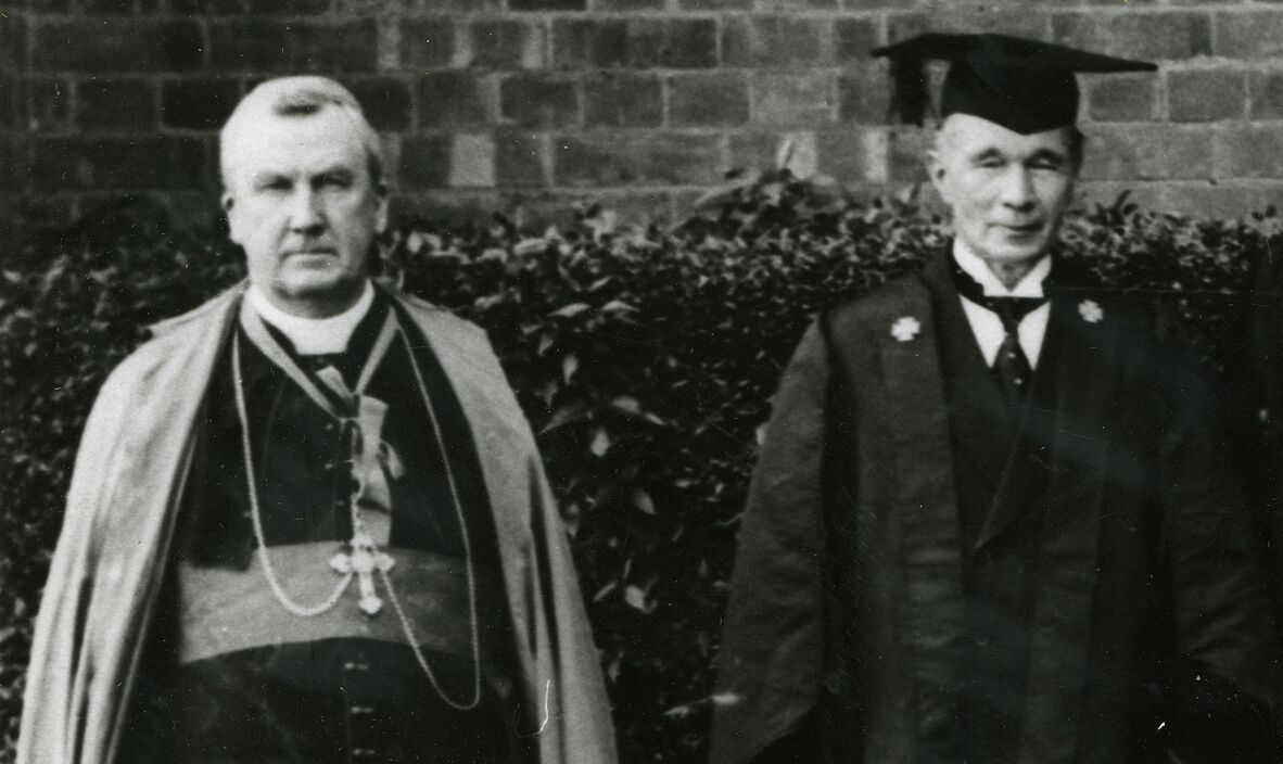Black-and-white historic photograph of two people standing outdoors in formal academic and ceremonial attire, with a brick wall and shrubs in the background.