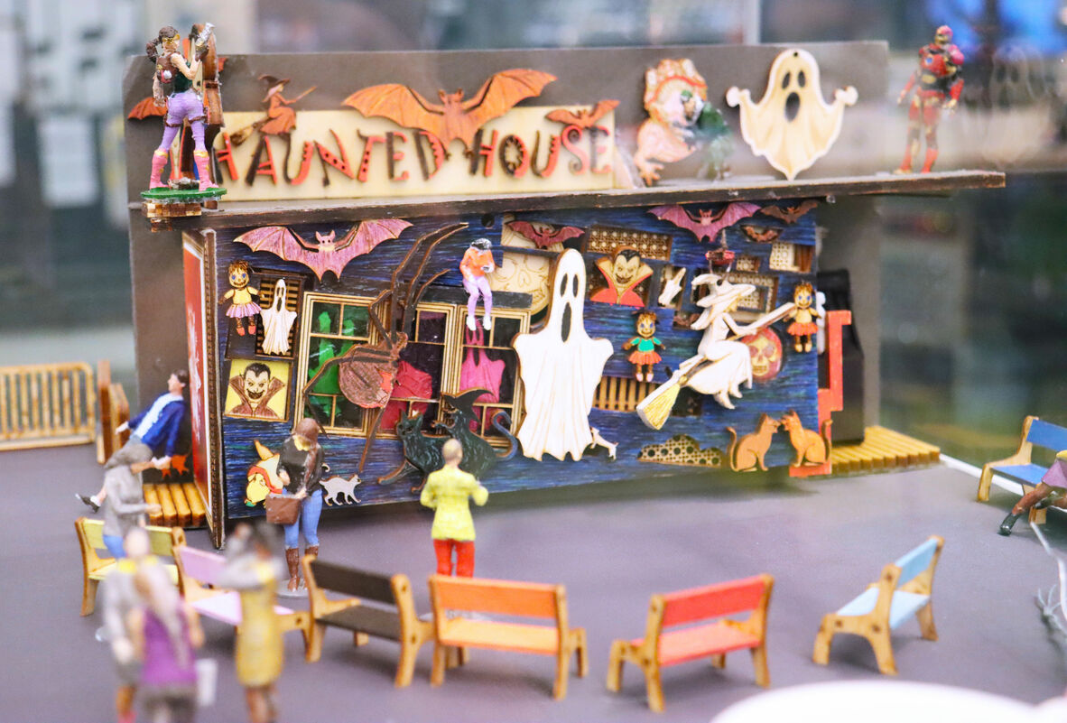 This is a miniature haunted house display with intricate details and a vintage horror theme. The facade is decorated with spooky elements such as ghosts, bats, witches, and classic Halloween characters. Above the building, a sign reads "Haunted House" in a stylized, eerie font. Small figures of people are arranged in front, observing the attraction, adding a sense of scale and realism. The display is vibrant, with colors like orange, purple, and green, creating a lively yet creepy atmosphere.