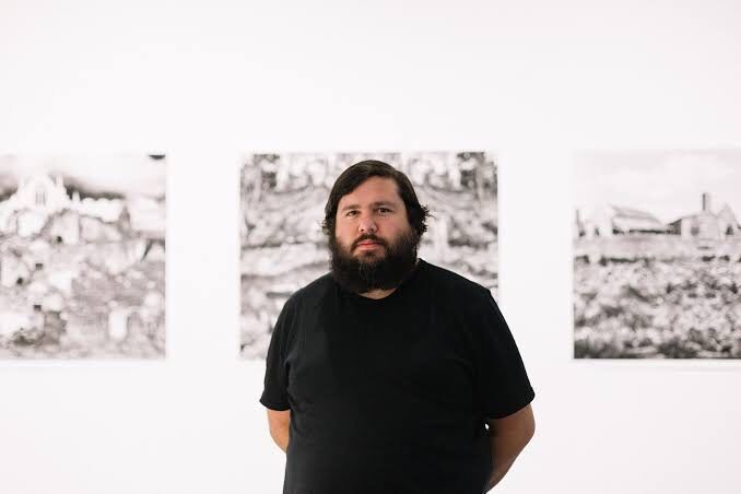 A man wearing a black t-shirt stands in front of three artworks in a gallery