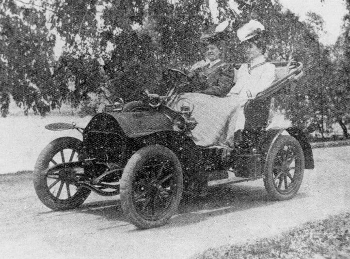 Two women sitting in an old car on a road surrounded by trees