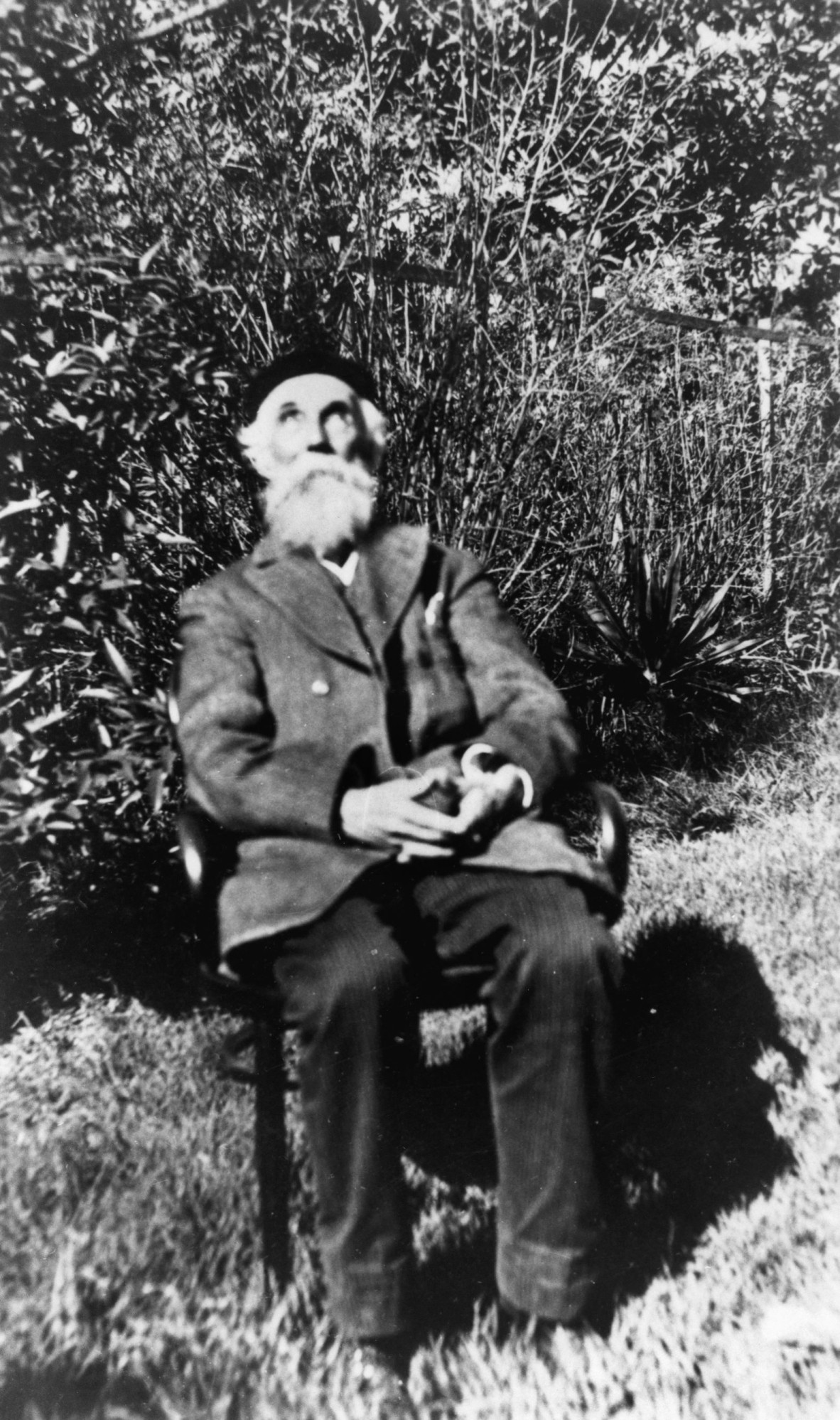 Black & white photo of an older man with a white beart sitting on a chair outside.