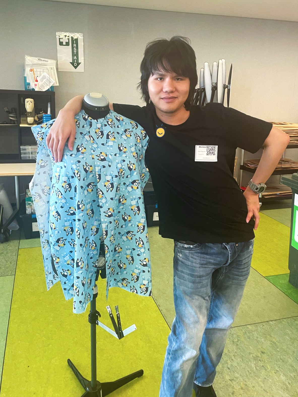 A photo of a young man next to a sewing mannequin with a shirt he is making
