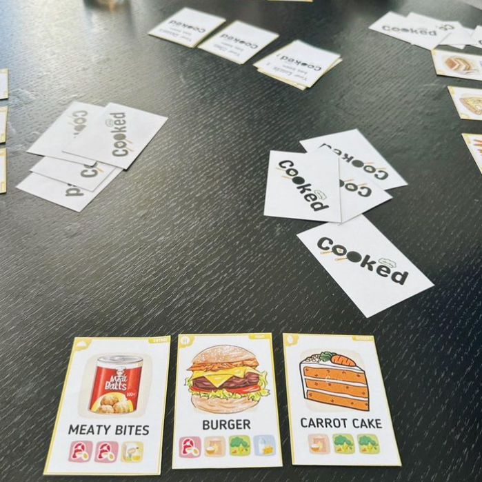 Close up photo of the Cooked board/card game of the cards. Shows an example of the graphics on the cards and the Early prototype versions.