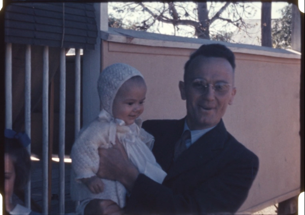 A still from a home movie, a middle-aged man in a dark suit smiles at the camera, holding a young child