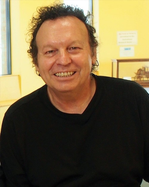 Headshot of Edoardo Crismani. He is seated at a desk with a large book in front of him. Edoardo wears a black jumper and is smiling