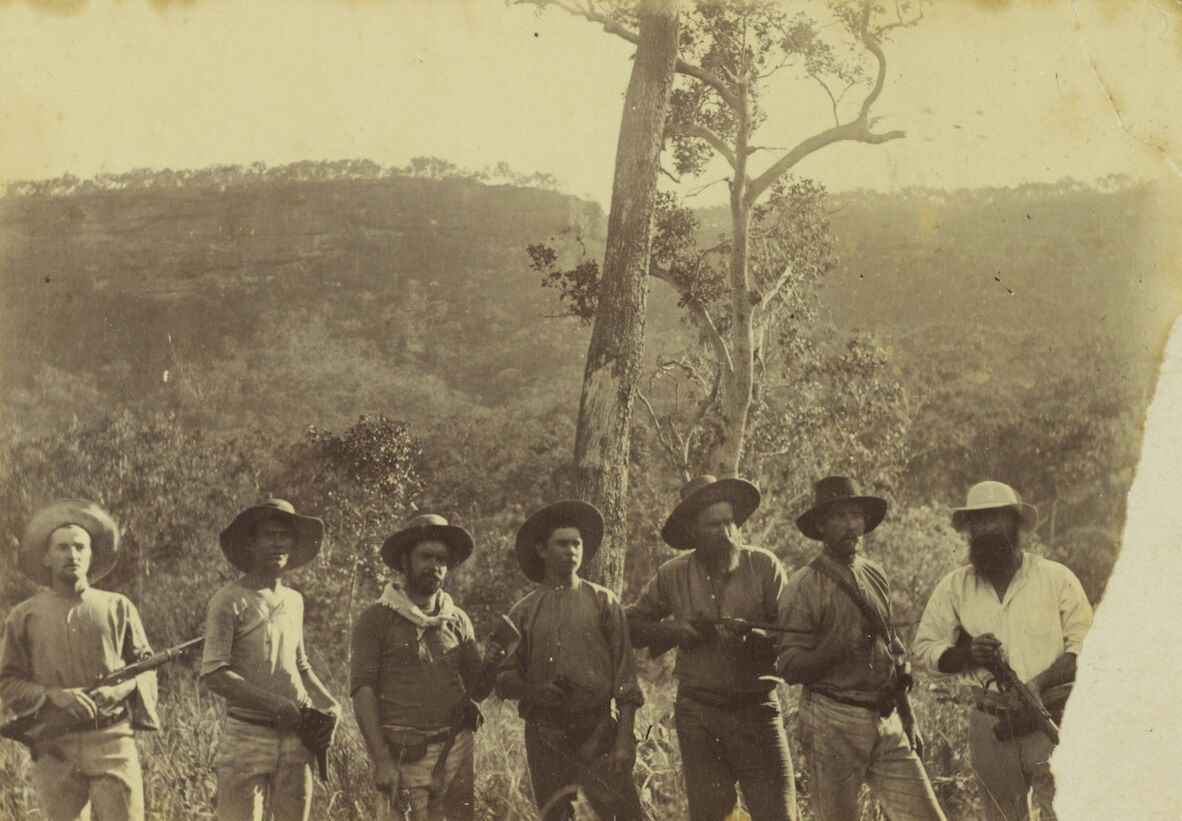 A black and white photo of 7 men in hats, armed with rifles, in the bush