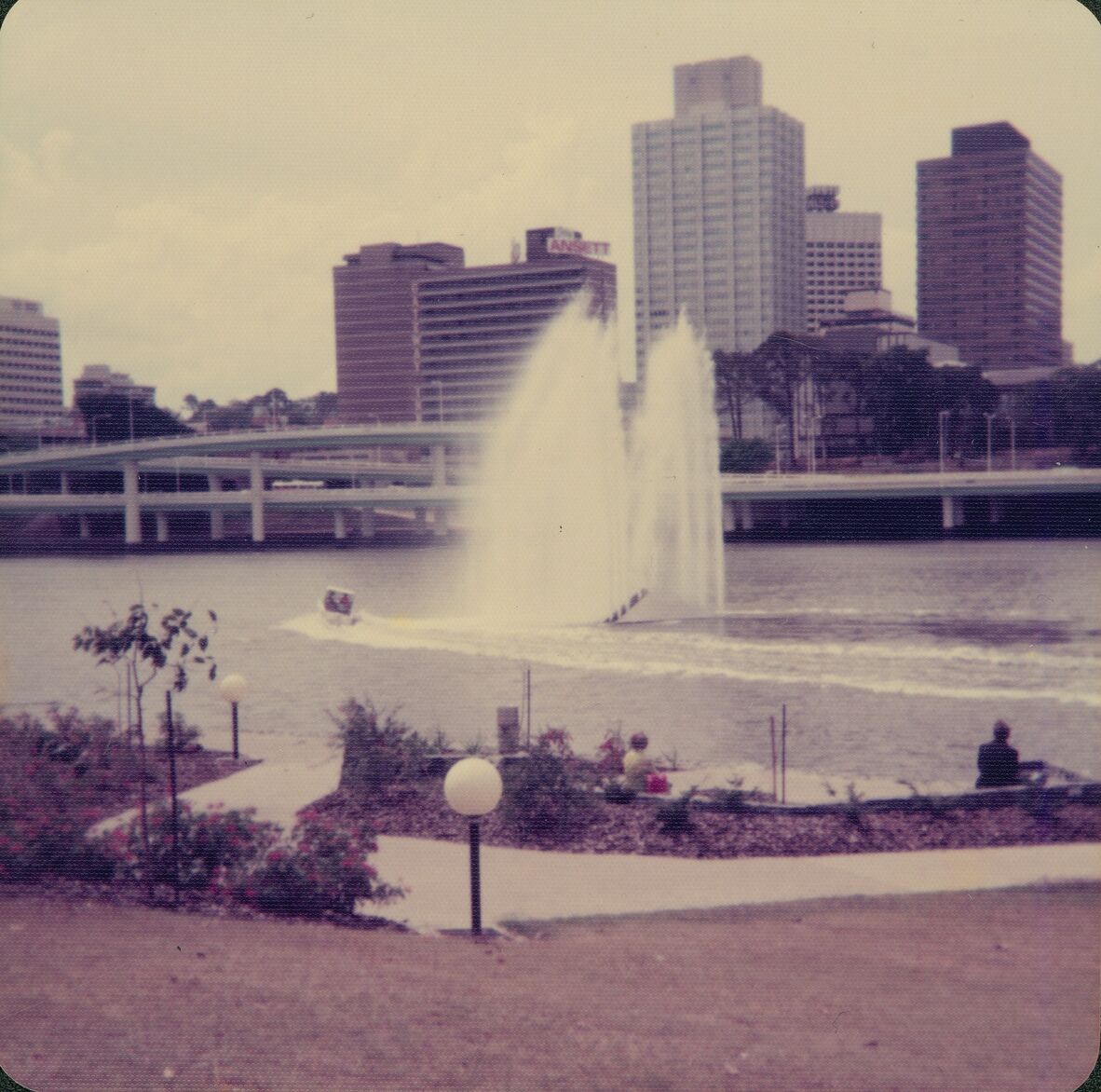 A sepia coloured photo of the Queen Elizabeth Silver Jubilee Fountain on the Brisbane River, Brisbane, Queensland, 1977. In the photo Brisbane city skyland can be seen in the background, with the founation's water reaching hige into the air.