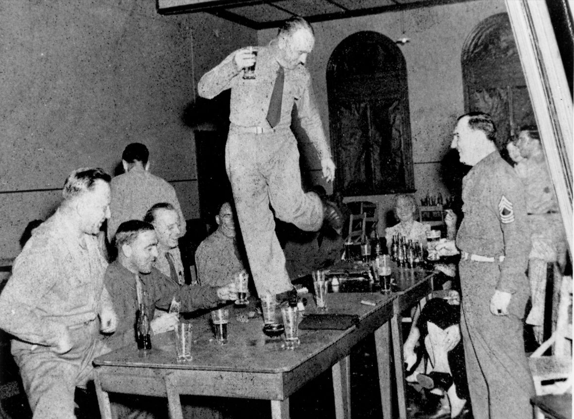 An archival, grainy black-and-white photograph shows a US soldier dancing on a long wooden table during a lively gathering in a high-ceilinged room. Balancing on his left leg with his right leg bent in mid-air, the soldier holds a glass aloft in his right hand. He wears a button-down shirt, a tie, and trousers.