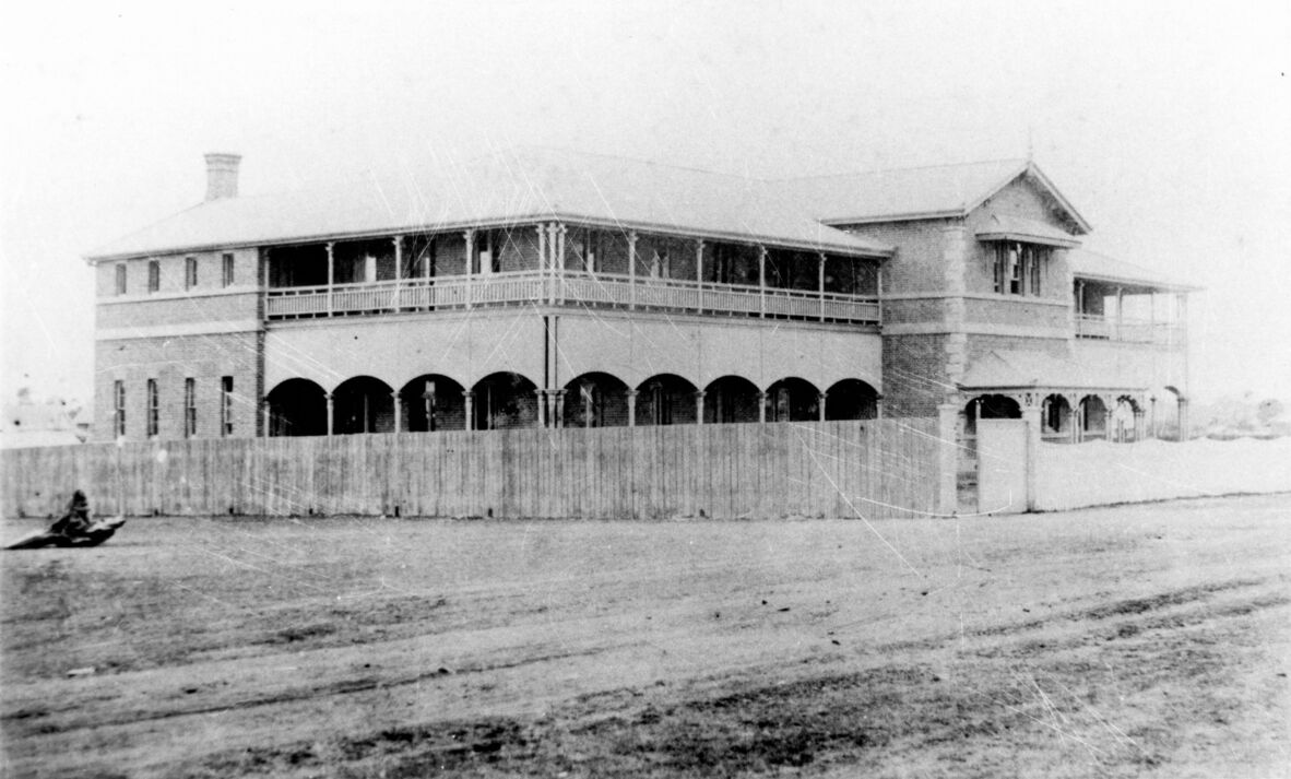 Black and white photo of two storey brick building with verandahs and a timber fence.