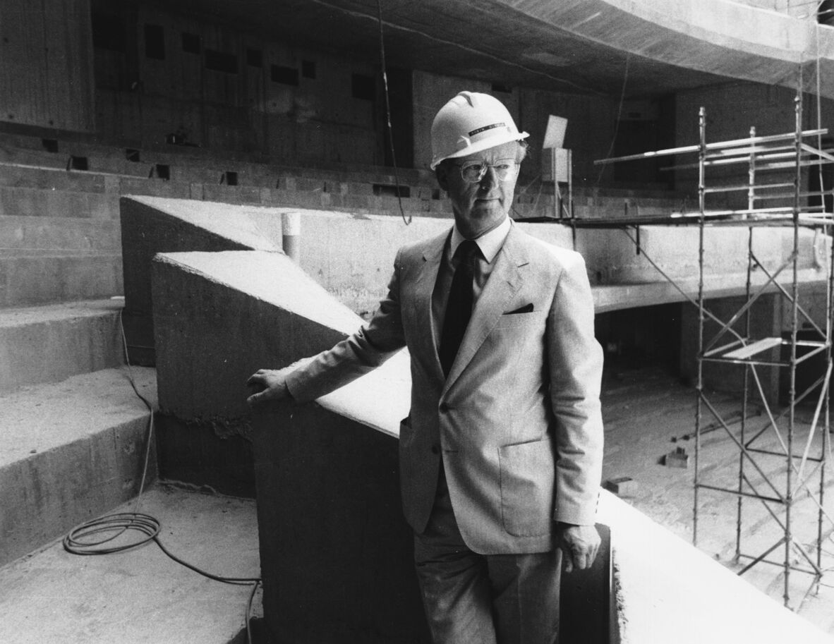 A black and white photo of Robin Gibson, architect of the Queensland Cultural Centre, standing on Balcony 2 of the Lyric Theatre of the Queensland Performing Arts Centre during construction. He is wearing a white hard helmet and is holding onto the side of the steps.