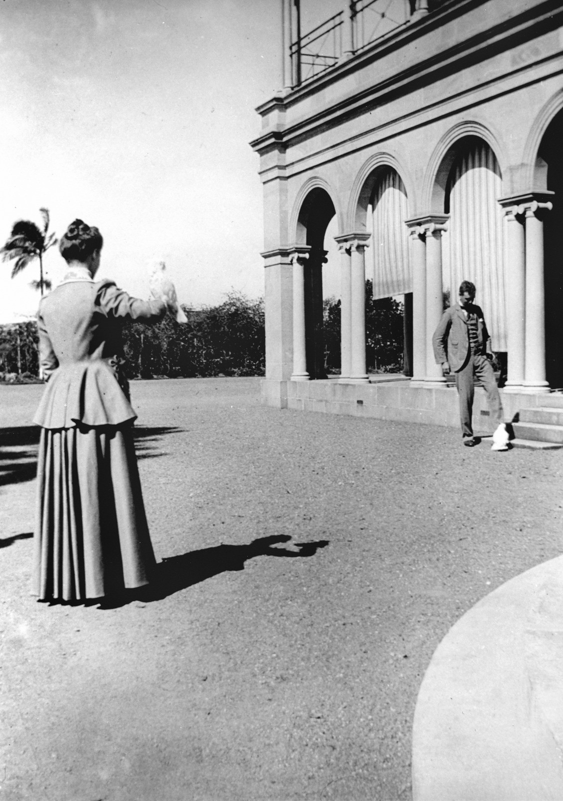 Black and white photo of a woman in a dress with her back to the camera with a bird perched on her hand. At right, a man in a suit looks at a white bird on the ground. The location is Government House, Brisbane.