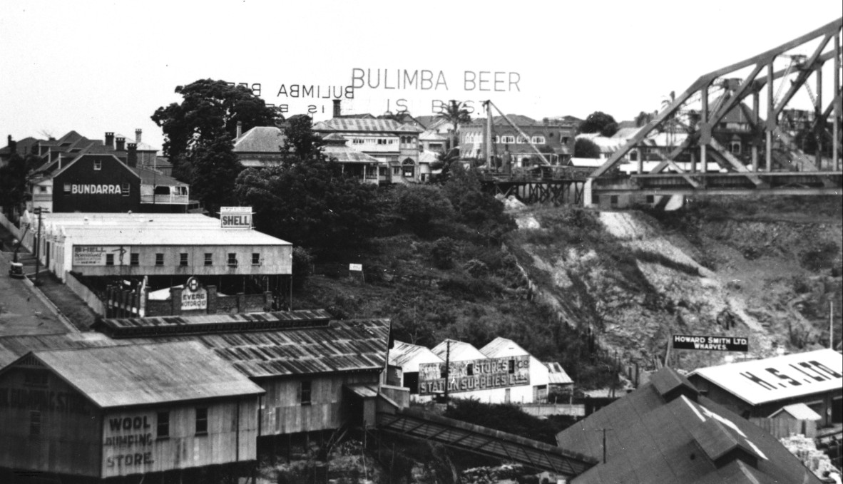 View of a section of the Story Bridge during construction. Buildings in Fortitude Valley, near the bridge's north entrance, can also be seen. The Bulimba brewery is prominent and the Howard Smith Ltd. wharves are visible in the bottom right corner of the photograph.