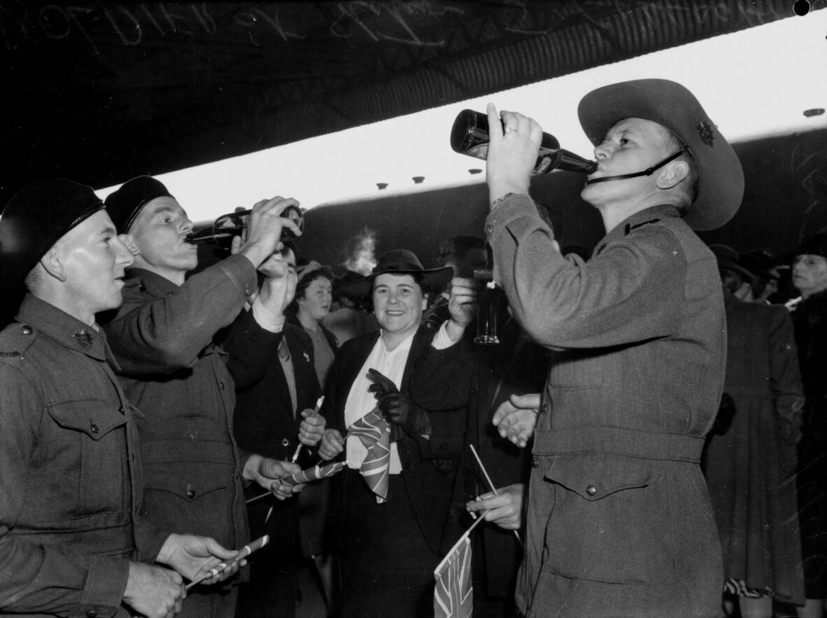 An archival black-and-white photograph shows World War II-era Australian soldiers in uniform celebrating, with two soldiers in the foreground drinking directly from bottles of beer. One on the right wears a traditional slouch hat, while the soldier on the left wears a side cap. Between them, civilians, including a smiling woman wearing a dark hat and coat, hold small Union Jack flags.