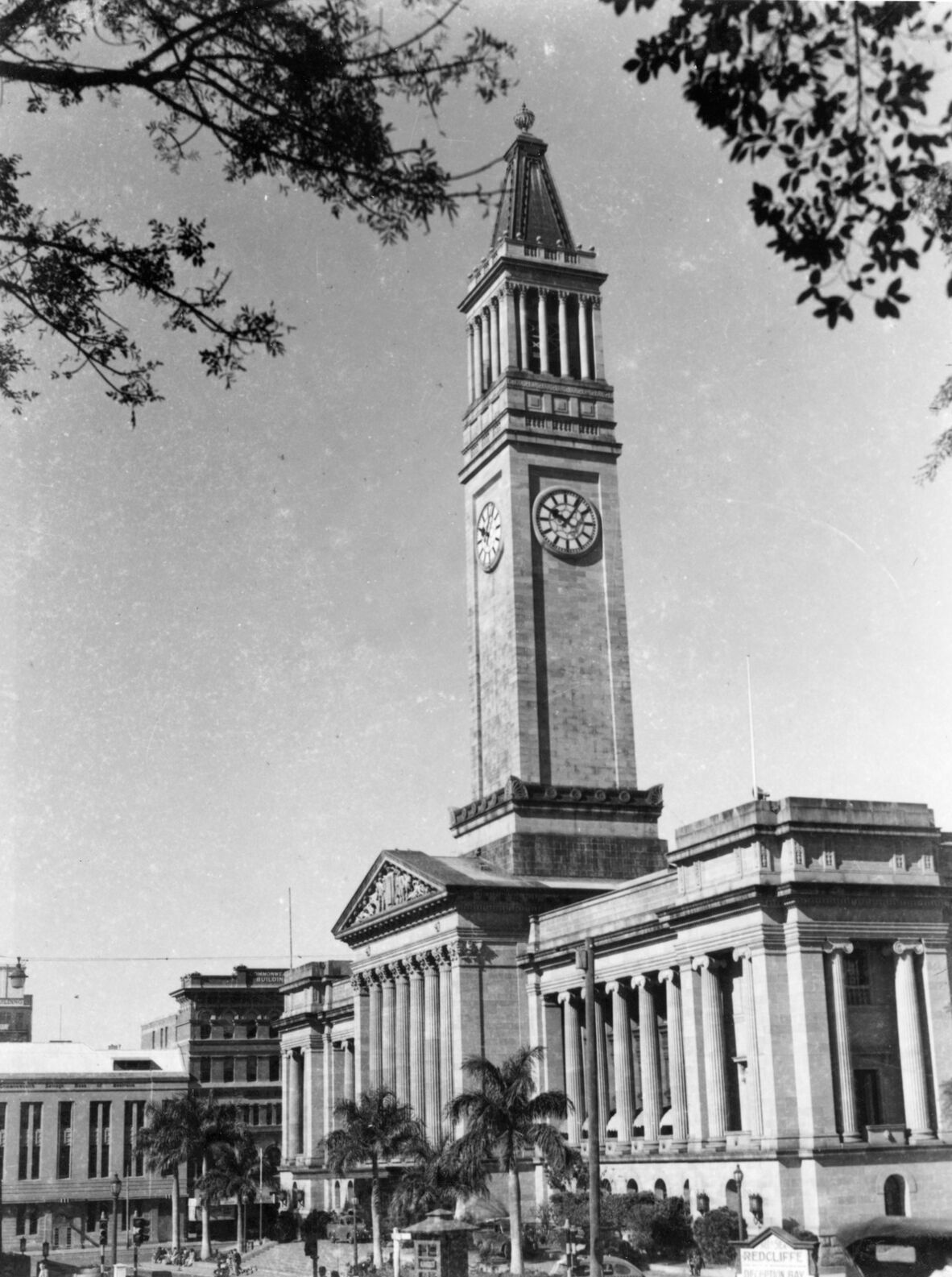Black-and-white image of Brisbane City Hall with its tall clock tower rising above classical columns, framed by leafy tree branches