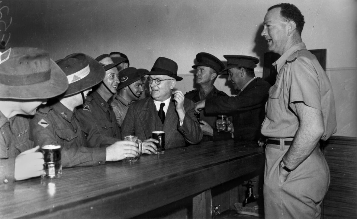 An archival, black-and-white photograph shows a group of men gathered at a wooden bar, smiling and socializing. Most of the men are dressed in military uniforms, including several wearing slouch hats. A man in the center wears a dark suit, a tie, glasses, and a fedora, holding a mug of beer. On the far right, a tall man in a short-sleeved military uniform stands with his hand in his pocket, facing the group. Mugs of beer sit on the long, polished wooden bar counter in the foreground.