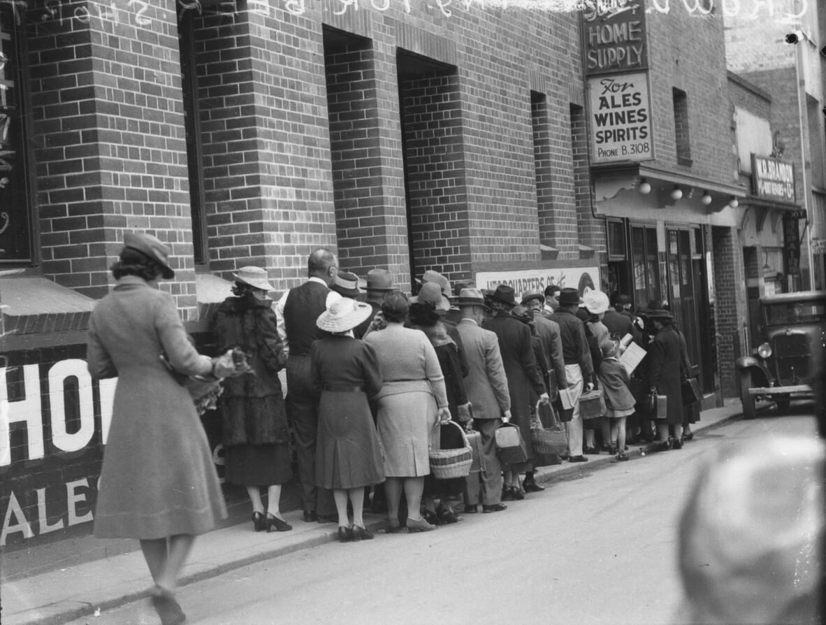 A black-and-white, eye-level shot captures a long queue of people on a sidewalk waiting to enter a building in the 1940s. A woman in a grey coat and a brimmed hat walks past the queue towards the left, holding a bouquet of flowers in her hand. Her back is partially to the camera, and she is captured in mid-stride. Behind her, a queue of around fifteen people, both men and women, stretches down the sidewalk. All of the men and women in the queue wear hats. Many of the women are in dresses or skirts and car