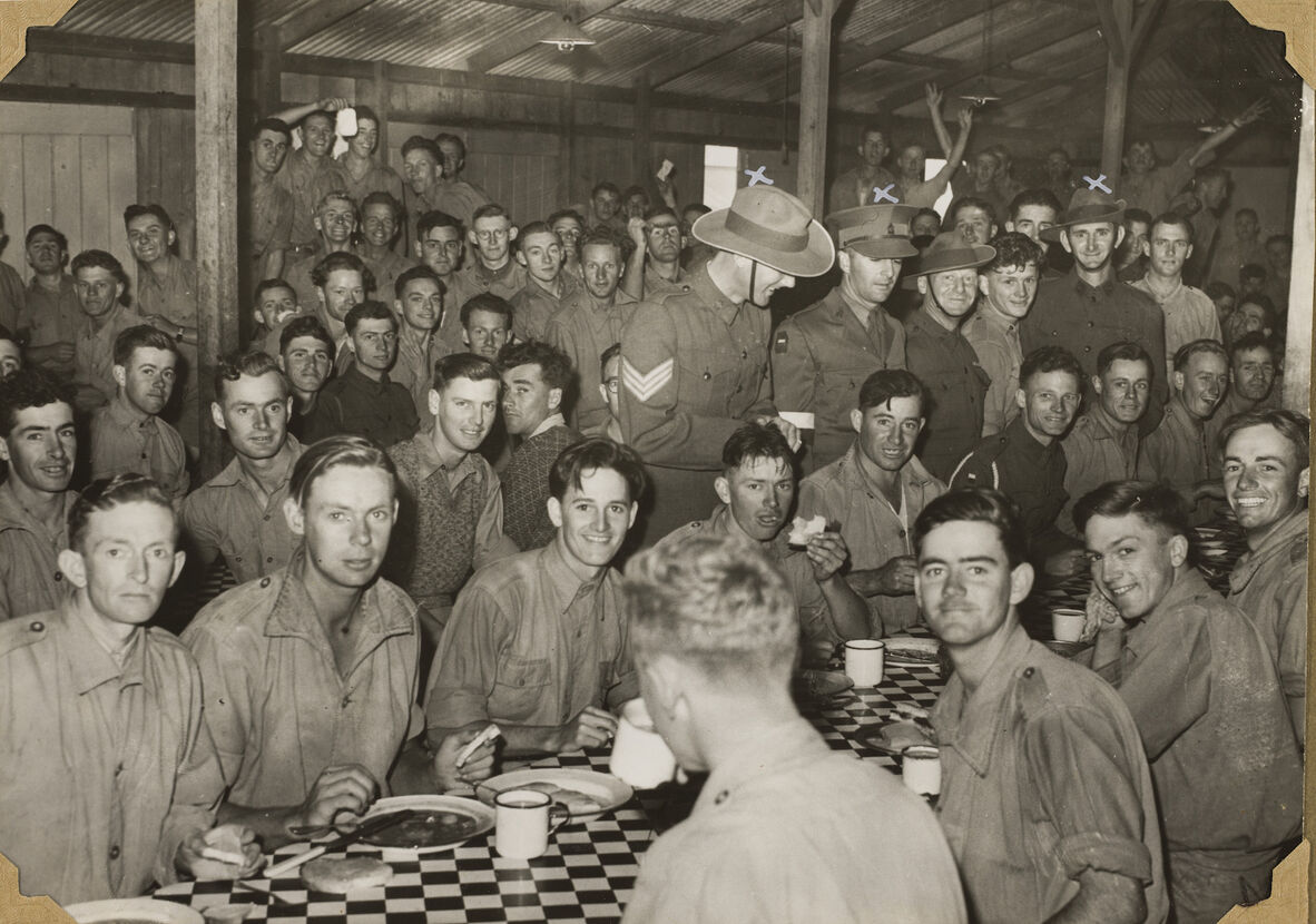 Black and white high-angle, medium-shot photograph of several Australian soldiers gathered in a mess hall around tables draped in a checkered tablecloth. The men are sitting and standing, with some smiling and looking directly at the camera. In the foreground, men sit at a long table with white mugs and plates. Standing behind them are other men, some wearing campaign hats and service caps. The space has wooden beams on the ceiling, with wooden walls in the background.