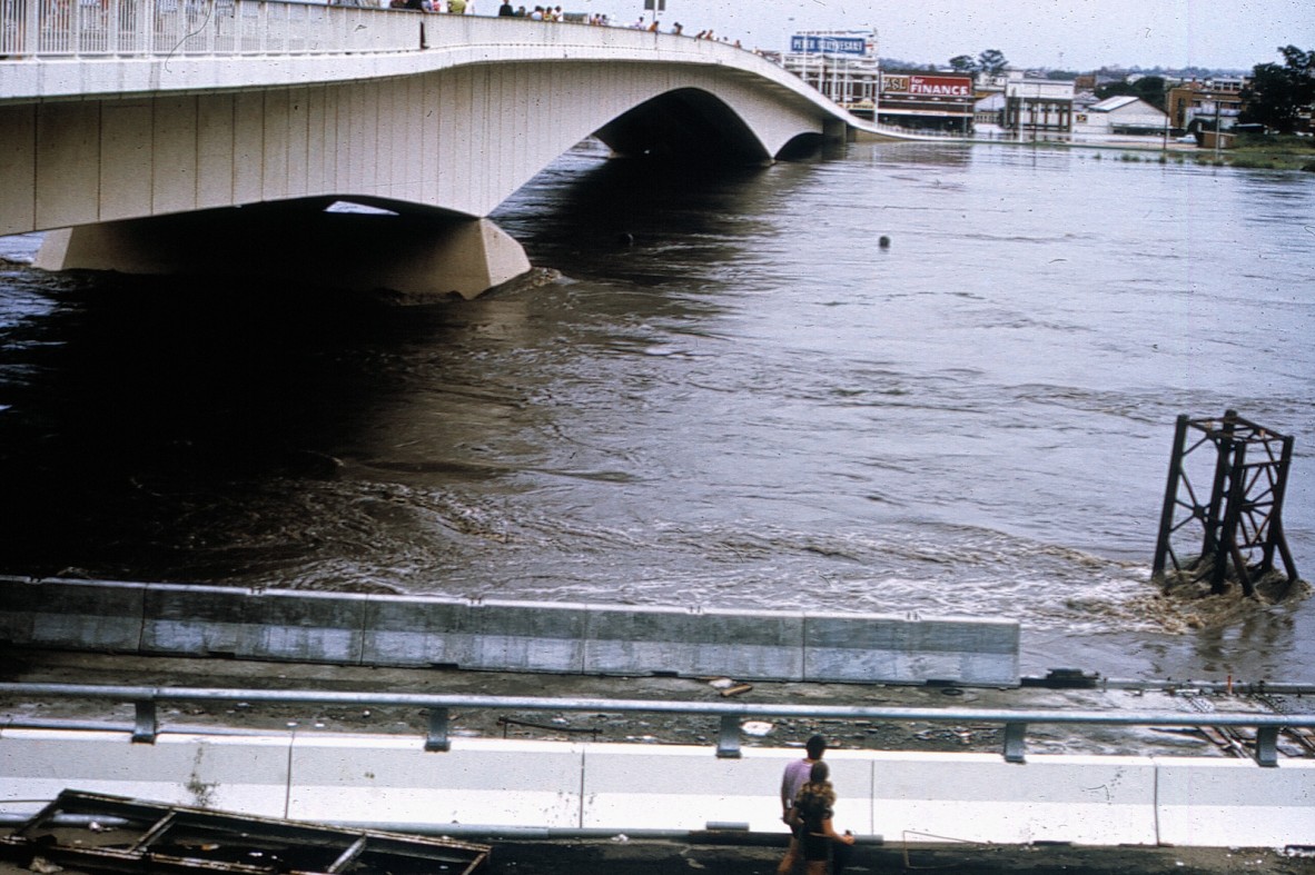 People are standing on the Victoria Bridge watching the fast flowing Brisbane River. The bridge road disappears under the water on the South Brisbane side of the city. 1974 Brisbane Flood