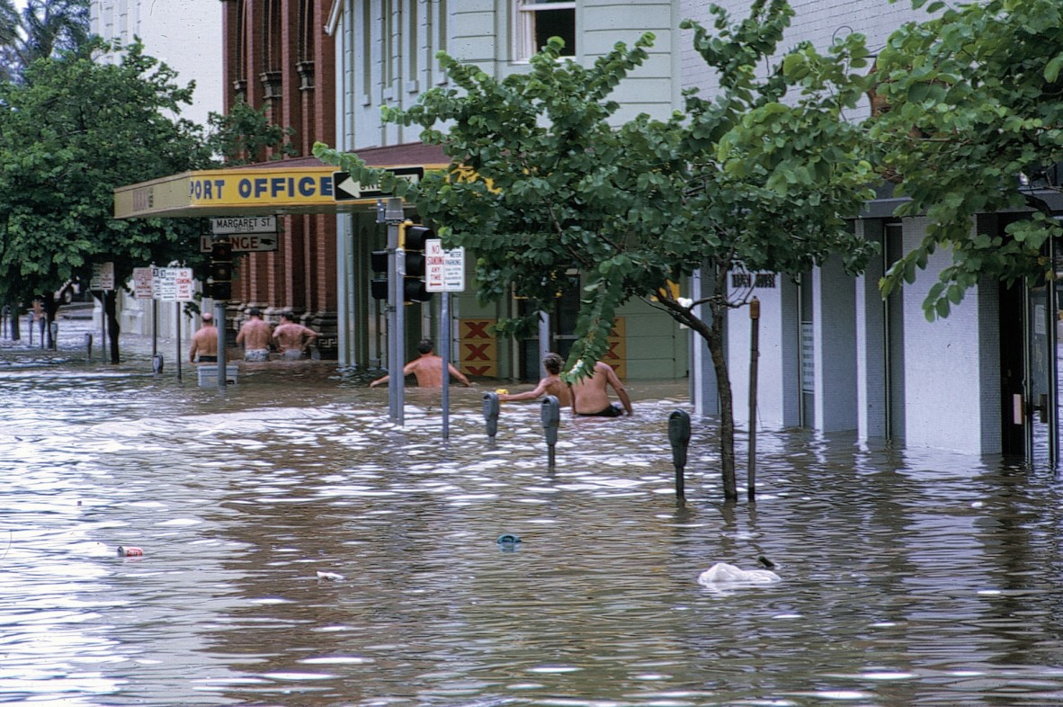  Men are wading waist deep in water towards the hotel and the parking meters in the street are almost submerged in some areas. A street sign for Margaret Street can be seen above the Lounge sign.