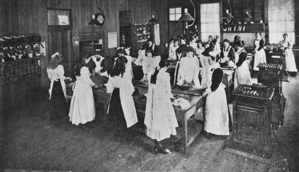 A black and white photograph of a group of women in aprons standing around benches, apparently in the midst of a cooking class.