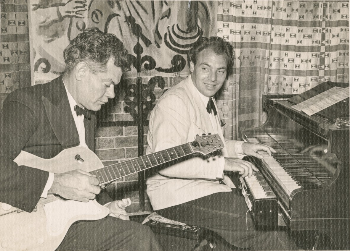 Darcy Kelly (left) playing his guitar seated next to a man playing a piano