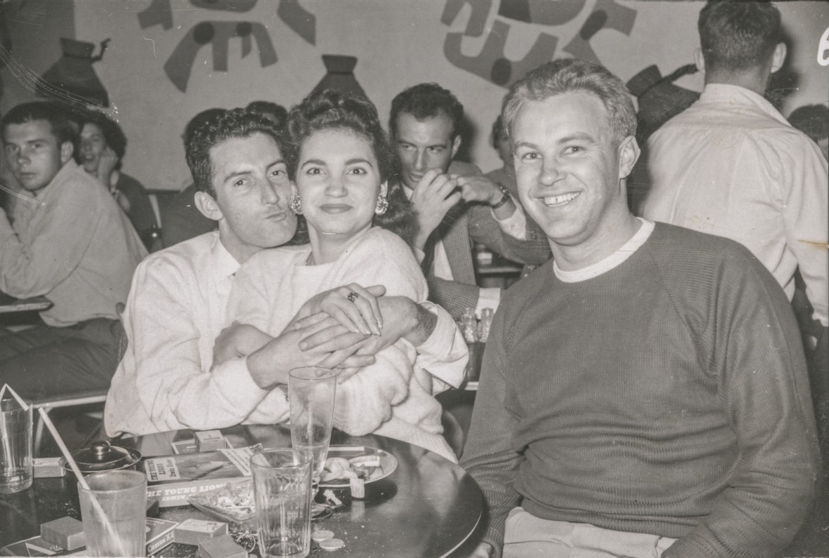 Three people sitting at a table with drinks and an ashtray