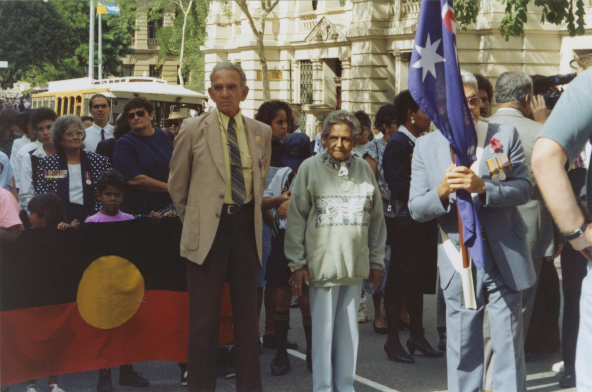 Len Waters and Oodgeroo Noonuccal standing in front of an Australian Aboriginal flag participating in a march, 1980.