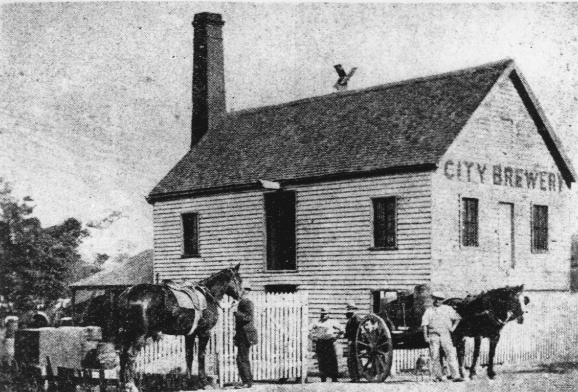 A black and white photographic print showing The City Brewery. Some workers and horses can be seen in the street in front of the two-storey, wooden building of the brewery. The chimney stack can be seen at one end of the building and the the signage at the front reads