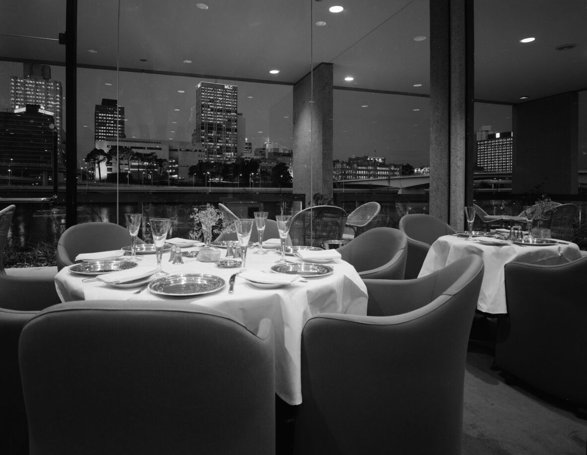 A black and white photo of some dinining round tables and chairs set up in front of a window overlooking the Brisbane skyline. The tables are draped in white tableclothes and can be seen to be set with plates, glassware and cutlery.