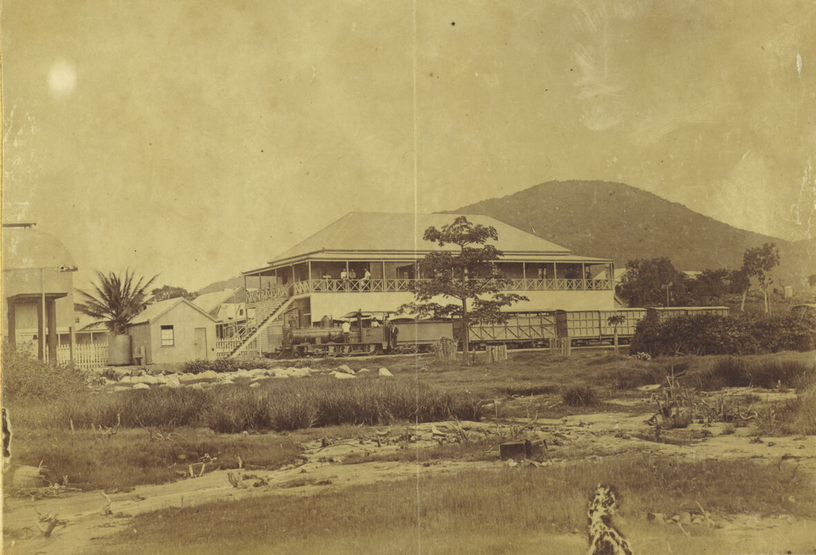 A faded black and white photo of a large building with a tin roof and a verandah. A mountain is in the background and there's scrub in the foreground.