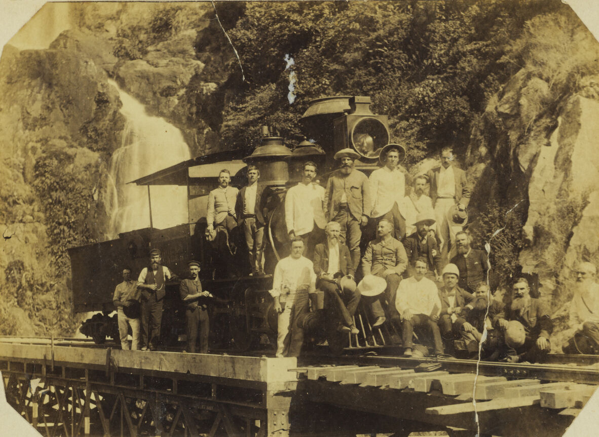 A black and white photo of people on a steam engine. Some are posing on the tracks beside a steep, rocky incline