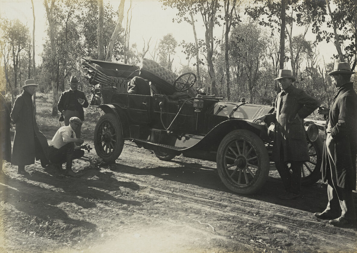 Black and white photo of six men standing around an old-fashioned motor car. One man is crouching and appears to be getting ready to fix a wheel.