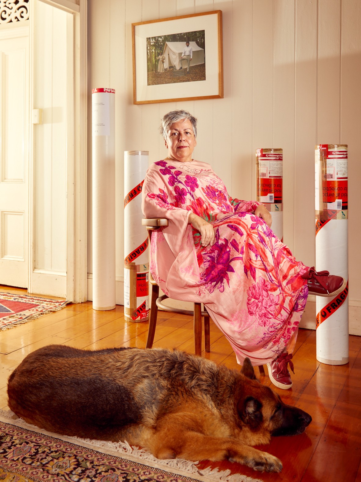 Artist Dr Fiona Foley in a pink floral dress seated in a Queenslander with white walls. A large dog rests at her feet and several large packing tubes for postage stand behind her.