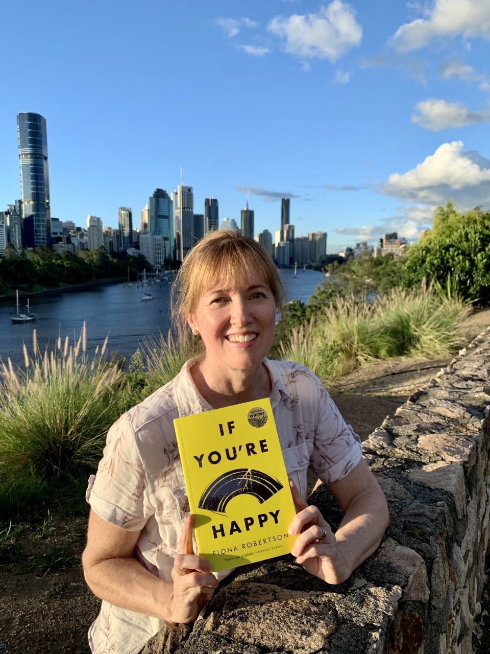 Fiona Robertson stands at Kangaroo Point cliffs holding a copy of her book The river and city of Brisbane are behind her