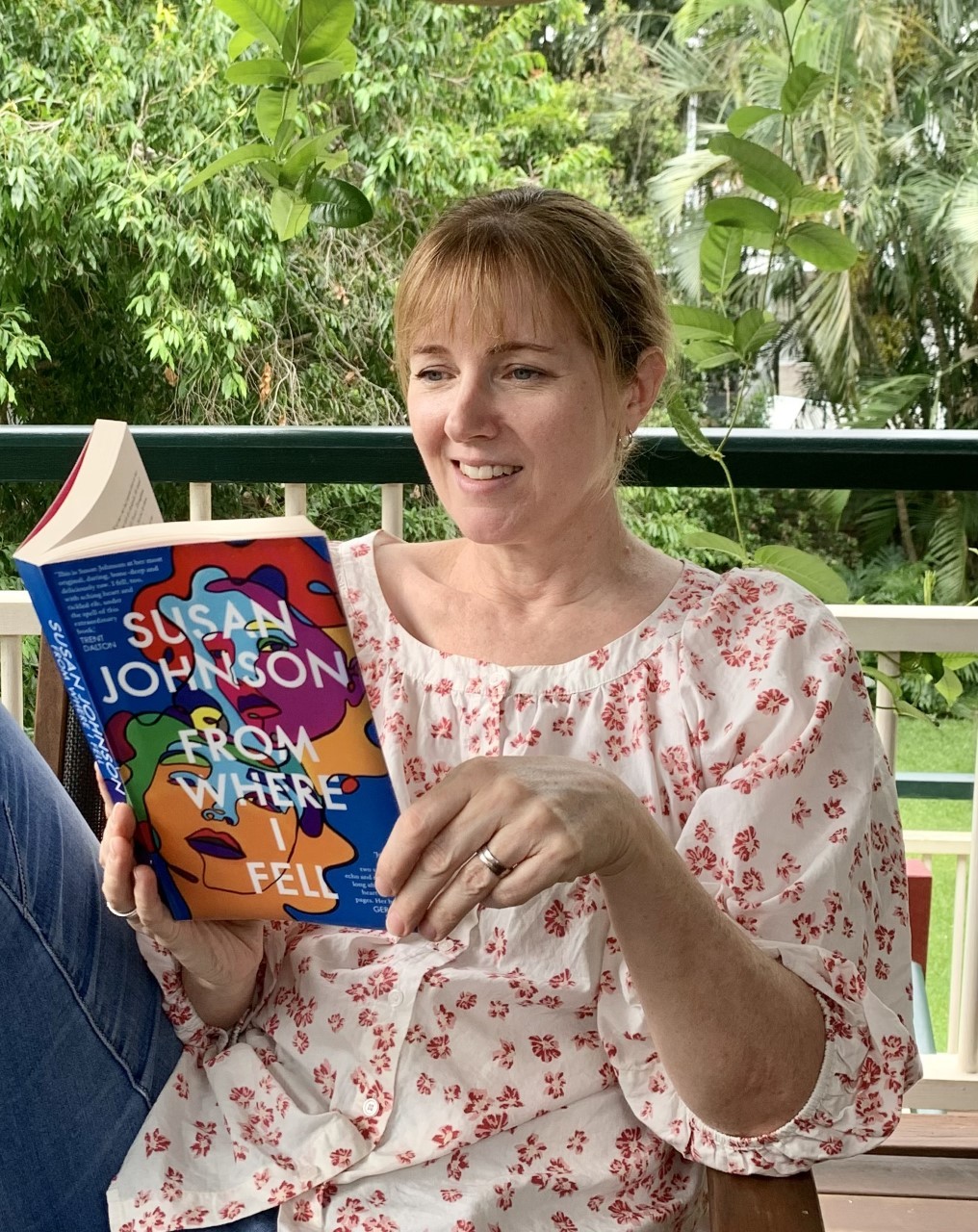 Fiona Robertson sits in a chair on her verandah. She is wearing a pink and white floral top and reading 'From where I fell' by Susan Johnsoning.