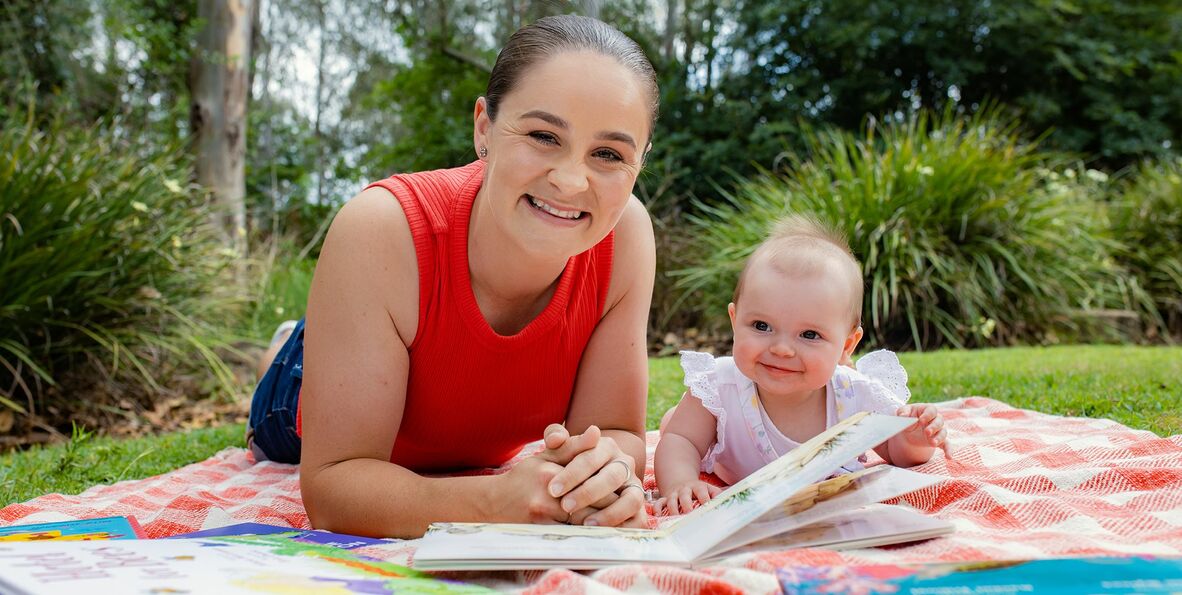 Ash Barty lying on picnic rug reading a book with a baby girl