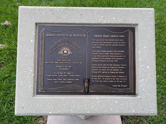 Small black plaque with text, designed to look like pages of an open book, set in white stone plaque, sitting upon grass.