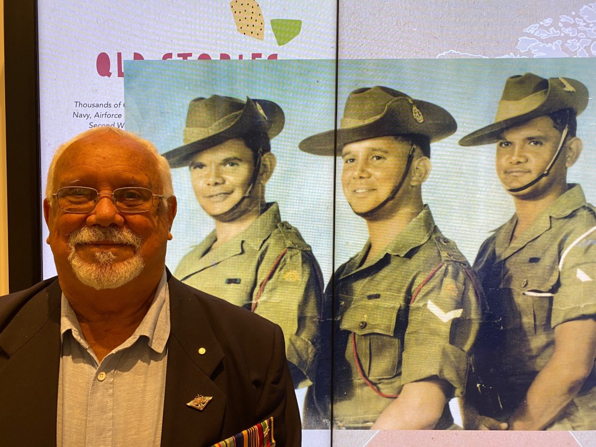 George Bostock standing in front of a enlarged photo of himself as a young soldier