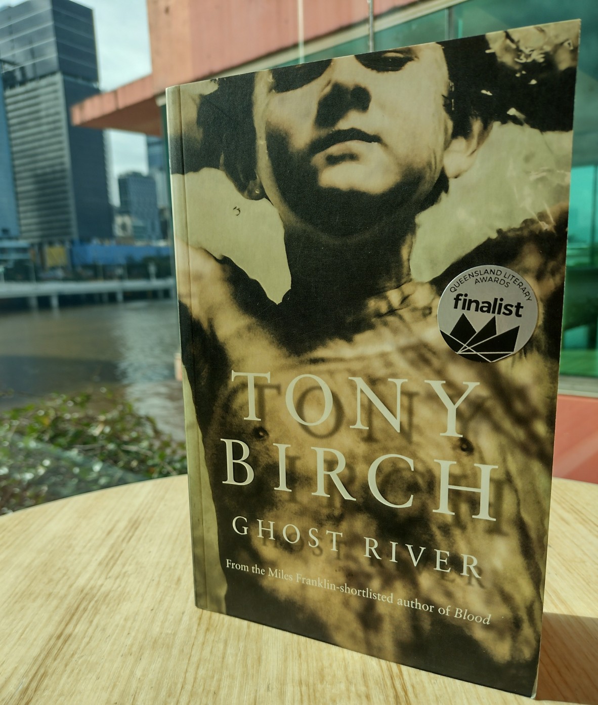A book sitting on a table in a light-filled space with a cityscape and glimpse of a river in the background The text on the cover reads Tony Birch Ghost River From the Miles Franklin-shortlisted author of Blood